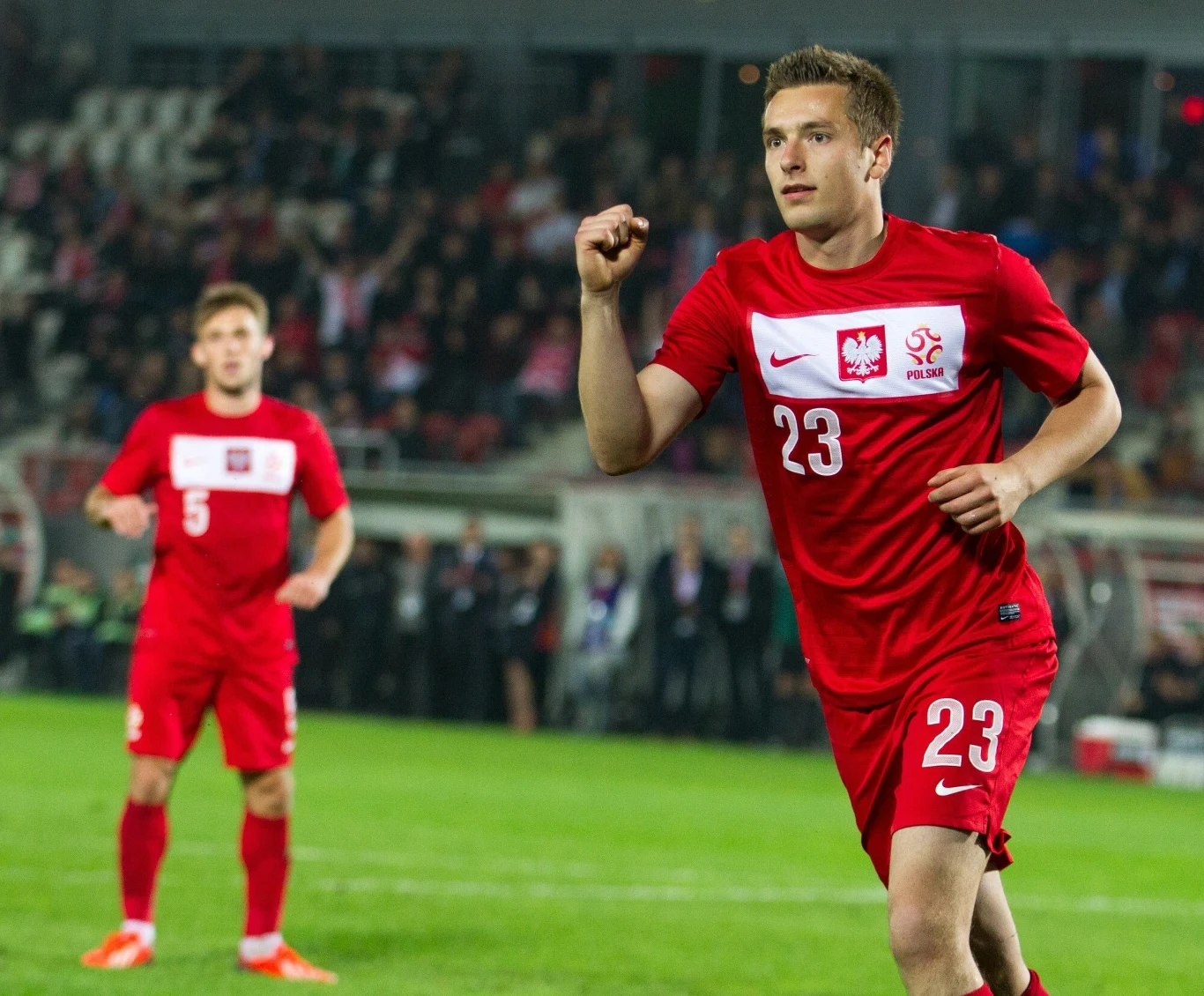 Piłkarz reprezentacji Polski w czerwonym stroju z numerem 23 świętuje zdobycie gola podczas meczu piłki nożnej, na drugim planie widać innego zawodnika w podobnym stroju oraz rozmytą publiczność na trybunach.