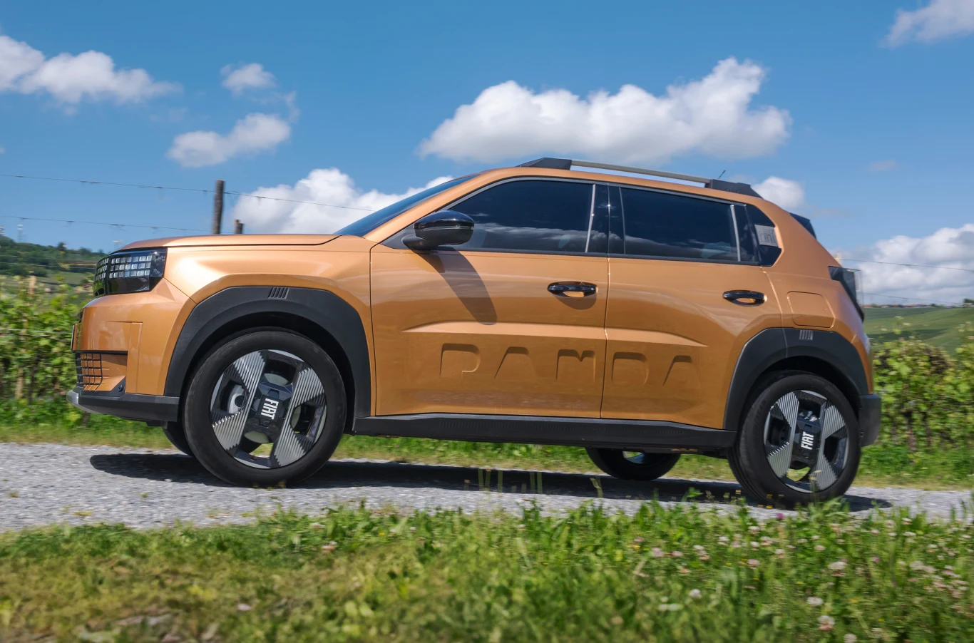 Nowoczesny SUV w kolorze pomarańczowym, zaparkowany na żwirowej drodze, w otoczeniu zielonej roślinności i pod jasnym, błękitnym niebem z drobnymi chmurami.