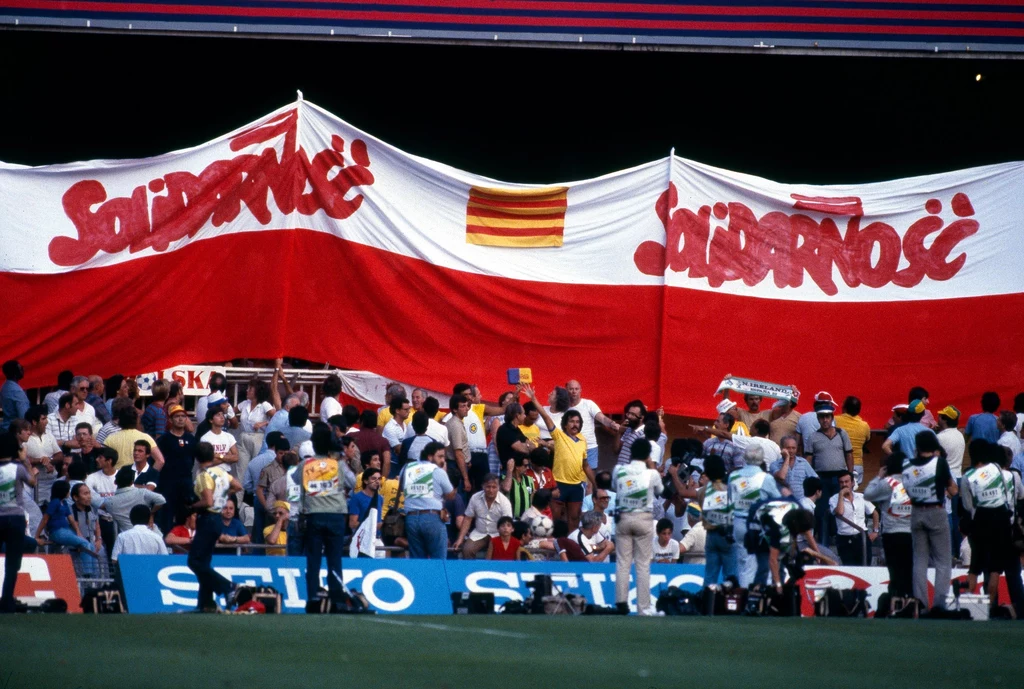"Solidarność" na trybunach Camp Nou Grupa ludzi zgromadzona pod wielką biało-czerwoną flagą z napisem 'Solidarność', w tłumie obecni fotoreporterzy i kibice, na pierwszym planie fragment boiska sportowego.