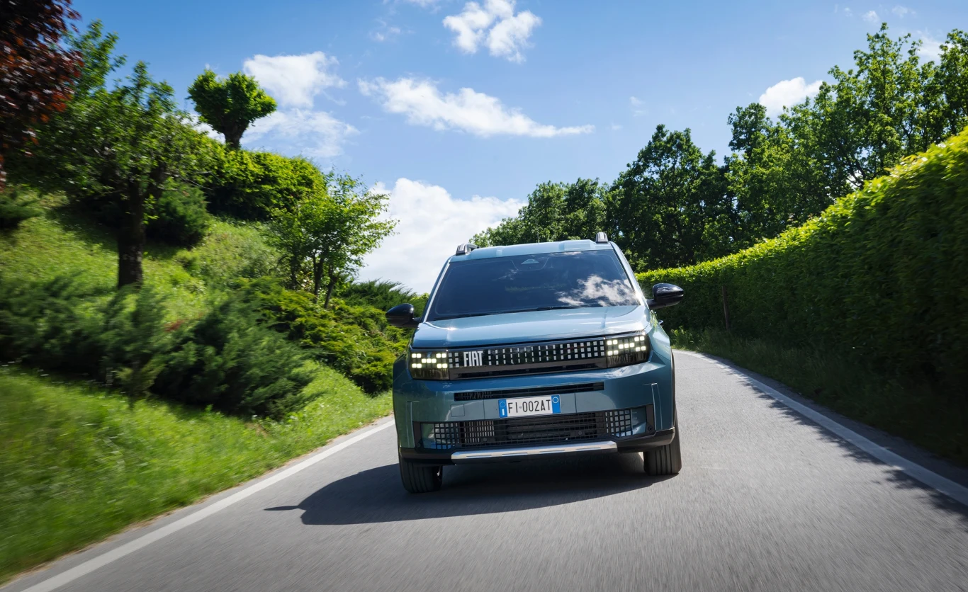 Samochód osobowy jedzie pustą asfaltową drogą otoczoną bujną zielenią, w tle widoczne są drzewa oraz błękitne niebo z chmurami.