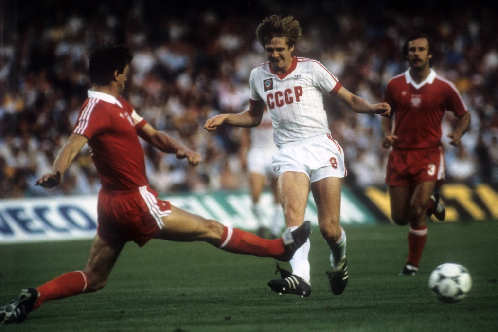 Mecz ZSRR - Polska, MŚ 1982 Dwóch piłkarzy w akcji na boisku podczas meczu, jeden z zawodników w białym stroju z napisem CCCP walczy o piłkę z rywalem w czerwonym stroju, w tle widoczny trybuny oraz inny zawodnik obserwujący sytuację.