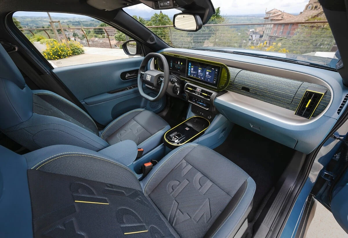 Nowoczesne wnętrze samochodu z cyfrową deską rozdzielczą, dużym ekranem dotykowym oraz elegancką tapicerką siedzeń utrzymaną w niebiesko-szarej kolorystyce, widok od strony pasażera i kierowcy, z widocznym krajobrazem przez okno.