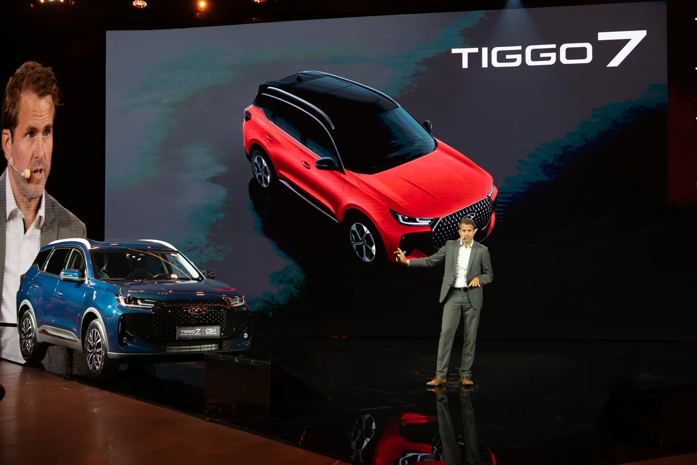 Duży ekran z wizualizacją czerwonego samochodu Tiggo 7, obok stoi niebieski model tego samego pojazdu; na scenie mężczyzna w garniturze prezentuje produkty, z lewej strony widoczna twarz innego mężczyzny w zbliżeniu.