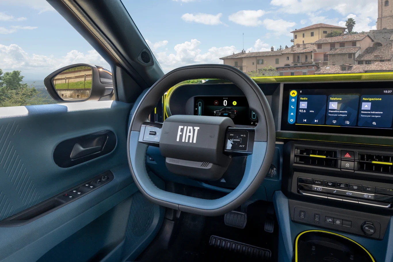 Nowoczesne wnętrze samochodu Fiat z cyfrowym kokpitem, kierownicą i dużym wyświetlaczem multimedialnym, przez okno widać zabudowania starego miasta i krajobraz z niebem oraz chmurami.
