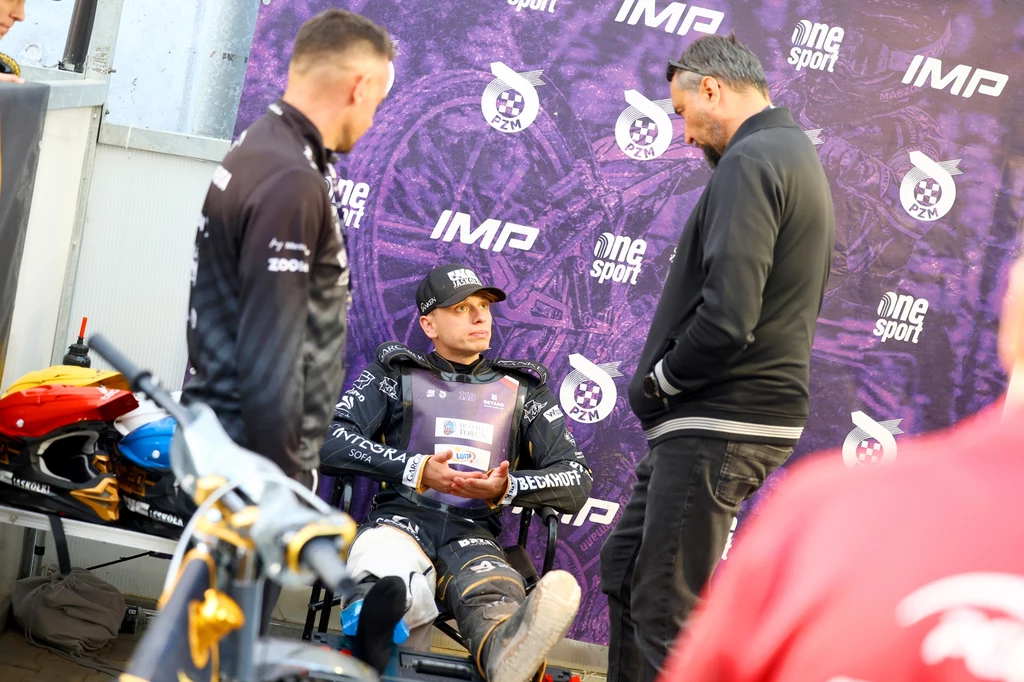 Jakub Krawczyk Zawodnik sportów motorowych w stroju wyścigowym siedzi na krześle, trzymając dyplom lub nagrodę; obok niego stoją dwaj mężczyźni rozmawiający z nim, w tle baner z logotypami wydarzenia.