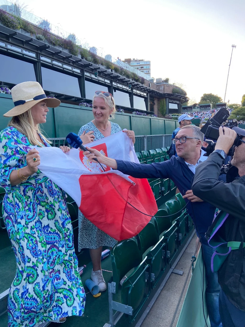 Dwie uśmiechnięte kobiety na trybunach stadionu trzymają polską flagę, wokół nich kilku dziennikarzy z mikrofonami i aparatami fotograficznymi, jedna z kobiet udziela wywiadu.