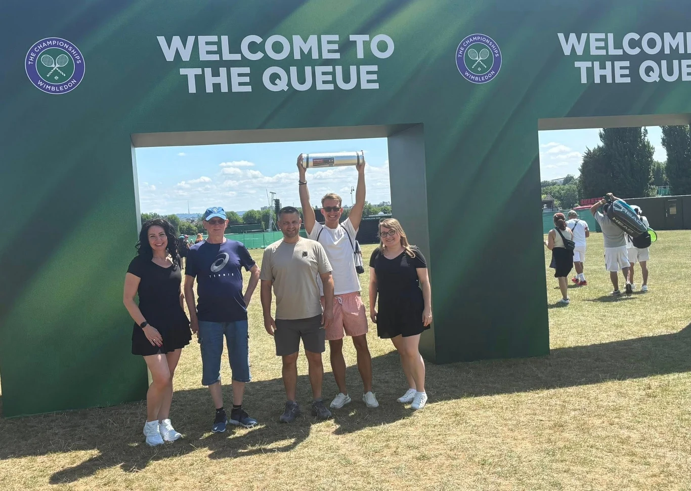 Grupa pięciu osób pozuje pod zielonymi bramami z napisem 'Welcome to the Queue', jedna osoba trzyma w górze przezroczysty pojemnik, wokół nich jasny trawnik oraz niewielki ruch innych osób w tle.