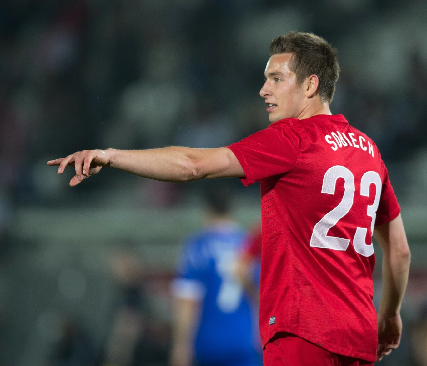 Piłkarz w czerwonym stroju z numerem 23 i napisem "Sukech" wykonuje gest wskazujący palcem, stojąc na boisku podczas meczu piłkarskiego, na drugim planie widoczny rozmazany zawodnik w niebieskim stroju.