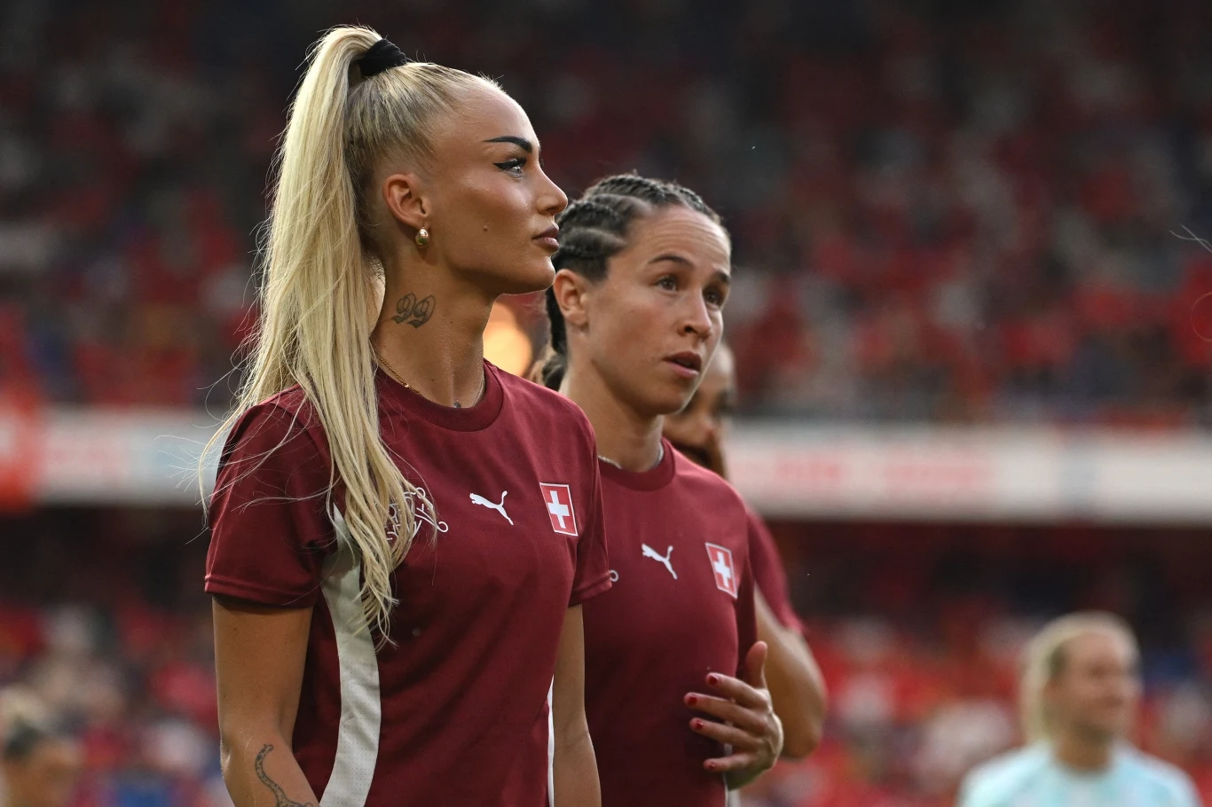Zawodniczki reprezentacji Szwajcarii w bordowych koszulkach z białym krzyżem, stojące na murawie stadionu, skupione przed rozpoczęciem meczu, w tle rozmyta publiczność.
