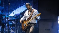 Muzyk grający na elektrycznej gitarze na scenie, w tle widoczna perkusja oraz kolumny wzmacniające, całość oświetlona klimatycznym światłem koncertowym.