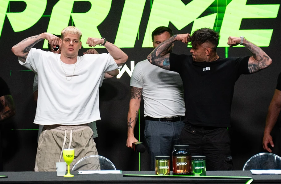 Prime MMA 13: "Taazy" vs Tańcula Dwóch mężczyzn na pierwszym planie napina mięśnie ramion, stojąc naprzeciwko siebie w pozie rywalizacyjnej, za nimi znajdują się inni uczestnicy wydarzenia, na stole widoczne są suplementy diety oraz napój.