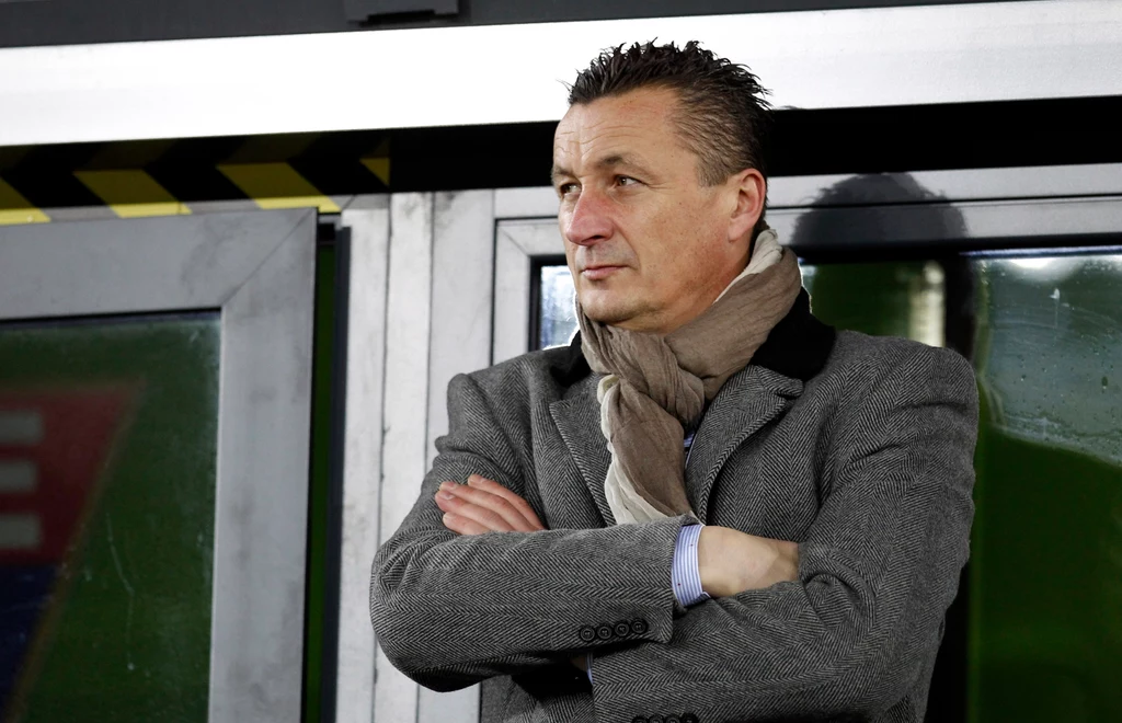 Tomasz Hajto Mężczyzna w średnim wieku ubrany w szary płaszcz i jasny szalik stoi z założonymi rękami, patrząc w bok, za nim znajduje się ściana i przeszklone drzwi z elementami ostrzegawczymi w żółto-czarne pasy.