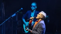 Wokalista śpiewający na scenie z mikrofonem w dłoni, w tle gitarzysta w ciemnych okularach. Atmosfera koncertowa, scena oświetlona punktowym światłem.