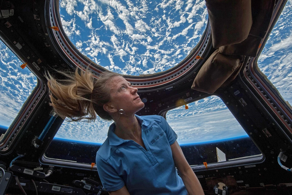 Astronautka NASA Karen Nyberg w module Cupola na Międzynarodowej Stacji Kosmicznej. Astronautka w niebieskiej koszuli dryfuje w module obserwacyjnym Międzynarodowej Stacji Kosmicznej, patrząc przez duży okrągły iluminator na Ziemię pokrytą białymi chmurami.