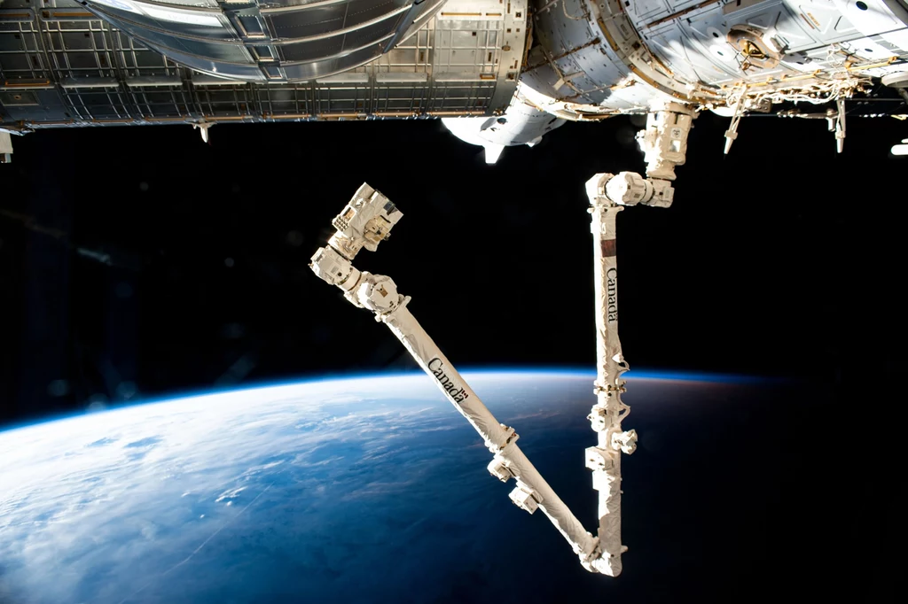Kanadyjskie ramię Canadarm2. Mechaniczne ramię stacji kosmicznej unoszące się nad Ziemią z widoczną atmosferą planety i fragmentem konstrukcji orbitalnej.