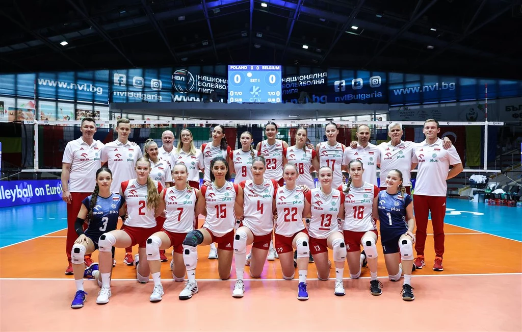 Reprezentacja Polski na ME U-20 w 2024 r. Drużyna siatkarek w pełnej obsadzie pozuje do wspólnej fotografii na hali sportowej, wszystkie zawodniczki i sztab ubrani w stroje reprezentacyjne, w tle tablica wyników oraz siatka.