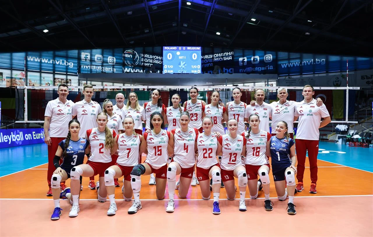 Reprezentacja Polski na ME U-20 w 2024 r. Drużyna siatkarek w pełnej obsadzie pozuje do wspólnej fotografii na hali sportowej, wszystkie zawodniczki i sztab ubrani w stroje reprezentacyjne, w tle tablica wyników oraz siatka.