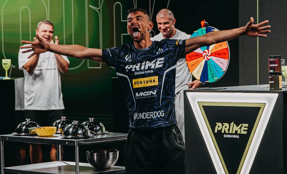 Mężczyzna w sportowej koszulce stoi z rozłożonymi ramionami i szeroko otwartymi ustami, wyrażając silną emocję, w tle widoczna grupa ludzi oraz kolorowe koło losujące i stół z przykrytymi daniami