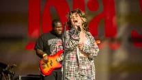 Wokalistka śpiewająca do mikrofonu na scenie ubrana w kraciasty strój, w tle gitarzysta basowy z czarnej koszulce i scenografia z rozmytym napisem. Klimat koncertu na żywo z widoczną energią i ekspresją artystów.