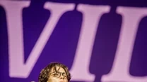 Młody mężczyzna śpiewający na scenie, trzyma mikrofon, ubrany w jasną koszulę i okulary, w tle wyraźne litery na dużym ekranie.