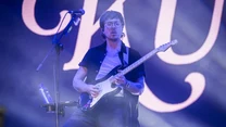Muzyk w okularach grający na gitarze elektrycznej podczas koncertu na scenie, w tle duże podświetlone litery i kolorowe światła.