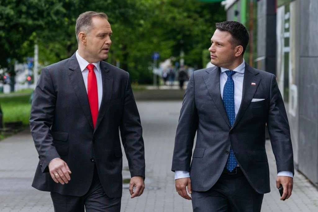 Prezydent elekt Sławomir Nawrocki i lider Konfederacji Sławomir Mentzen Dwóch elegancko ubranych mężczyzn idących chodnikiem obok budynku, rozmawiających ze sobą. Obaj mają na sobie garnitury, jeden z nich ma czerwony krawat, drugi niebieski. W tle widoczna zieleń oraz fragment ulicy.