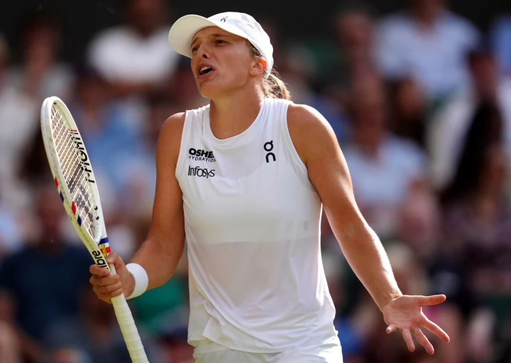 Iga Świątek w akcji podczas tegorocznej edycji Wimbledonu Tenisistka w białej sportowej koszulce i czapce prezentuje wyraz emocji, trzymając rakietę do tenisa w jednej dłoni i gestykulując drugą ręką, w tle rozmyta publiczność na trybunach.