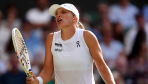 Tenisistka w białej sportowej koszulce i czapce prezentuje wyraz emocji, trzymając rakietę do tenisa w jednej dłoni i gestykulując drugą ręką, w tle rozmyta publiczność na trybunach.
