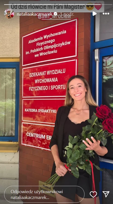 Natalia Bukowiecka obroniła pracę magisterską Uśmiechnięta młoda kobieta w eleganckim ubraniu trzymająca bukiet czerwonych róż na tle budynku Akademii Wychowania Fizycznego we Wrocławiu z widocznym szyldem informacyjnym uczelni.