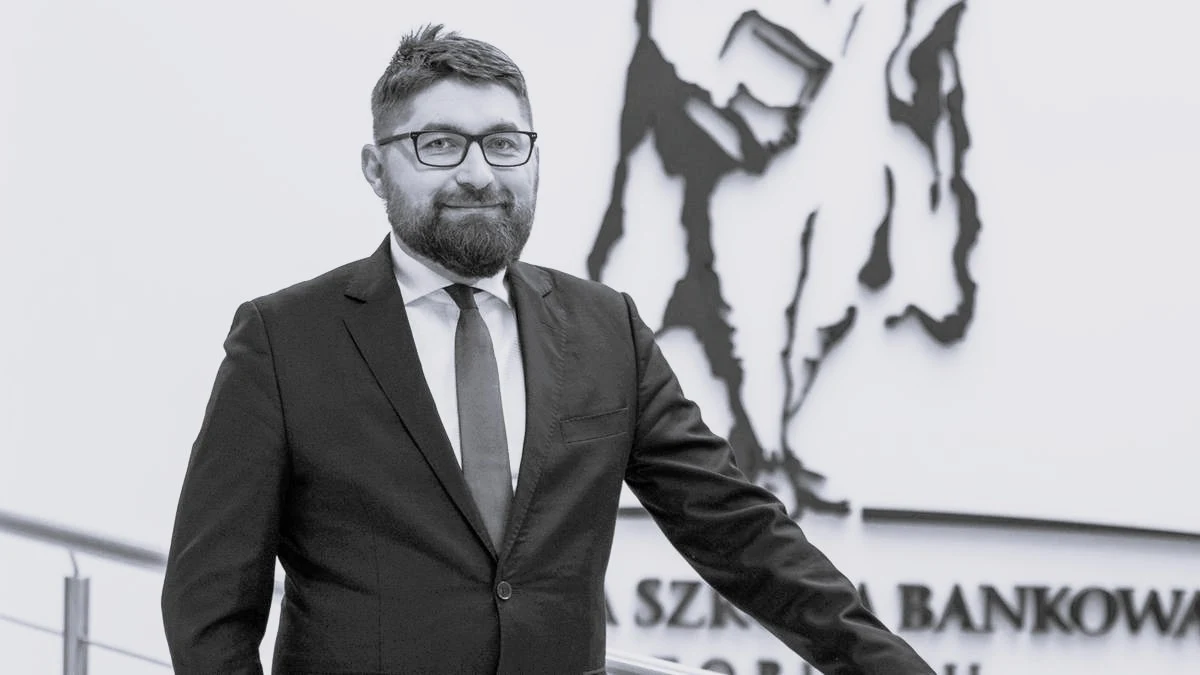 Uczelnia w Toruniu poinformowała o śmierci Jarosława Wojtasa Mężczyzna w garniturze z brodą i okularami stojący w eleganckim otoczeniu na tle napisu Szkoła Bankowa.