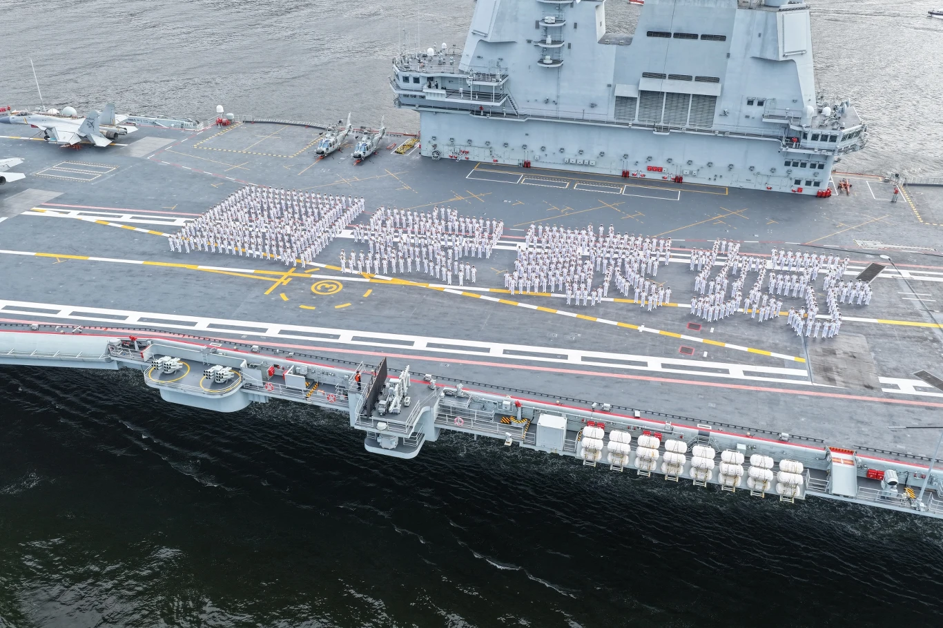 Zdjęcie z drona pokazuje oficerów i żołnierzy ustawionych w chińskie znaki "bezpieczny kraj, kochany dom" na pokładzie lotniskowca Shandong na wodach u wybrzeży Hongkongu Pokład lotniskowca z dużą grupą marynarzy w białych mundurach ustawionych w regularne formacje, tworzących napisy, w tle charakterystyczne nadbudówki i wyposażenie okrętu, otoczony wodą.