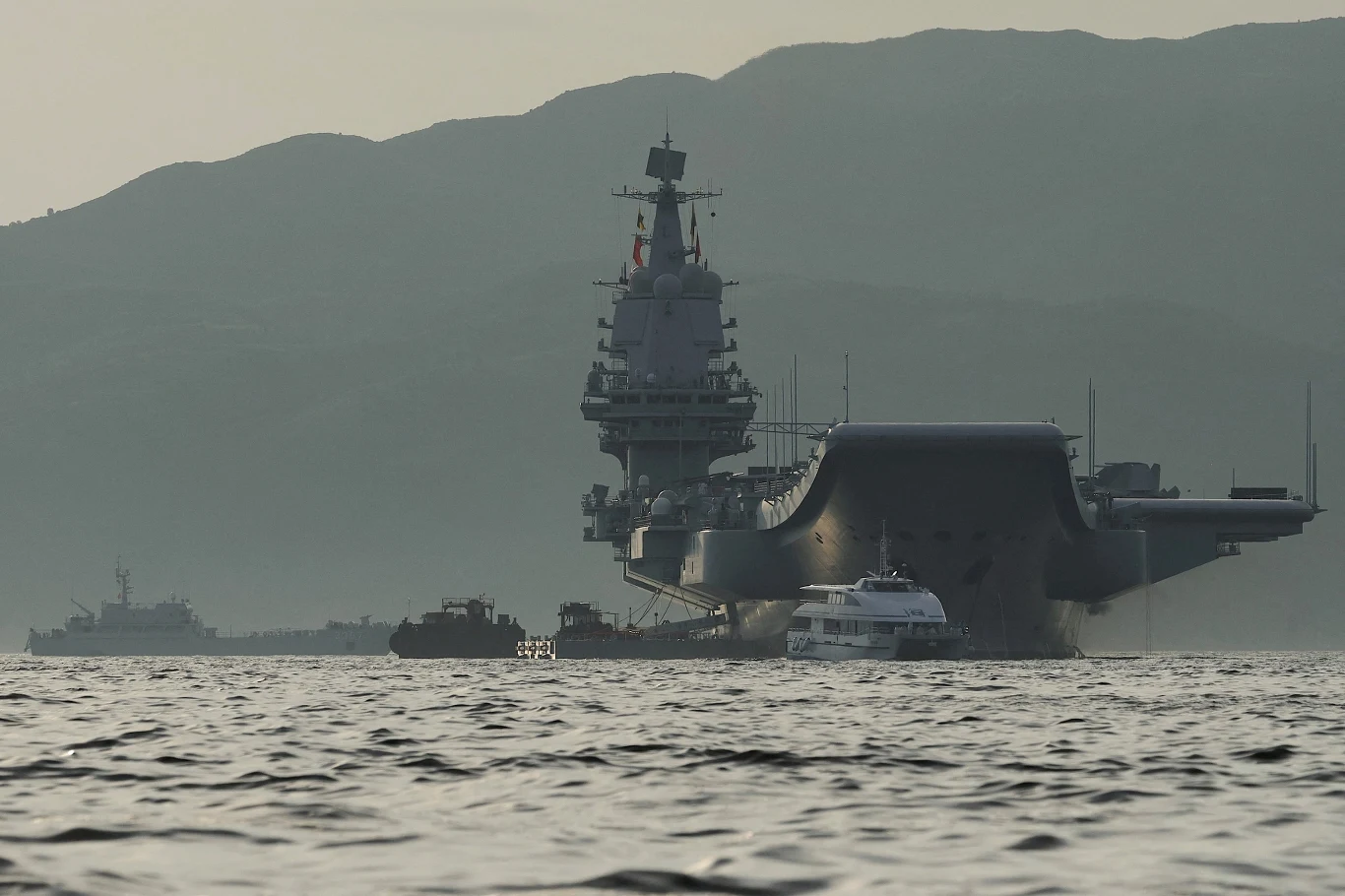 Lotniskowiec Shandong pływający w pobliżu Hongkongu Lotniskowiec wojenny płynący po spokojnej wodzie, otoczony mniejszymi jednostkami pływającymi na tle górzystego krajobrazu.