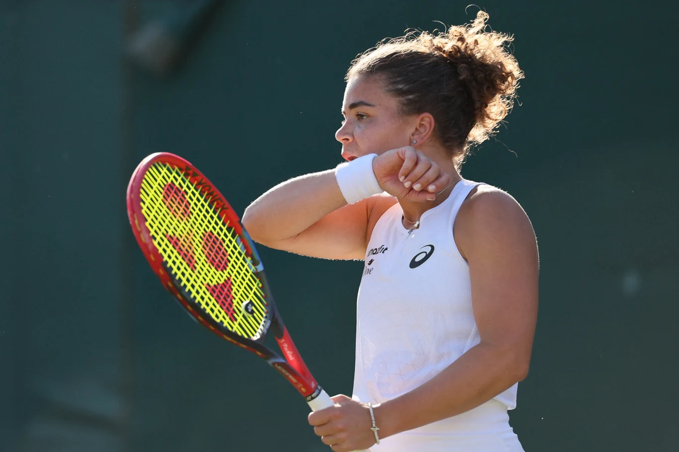 Jasmine Paolini najpierw odpadła z Wimbledonu w singlu, a teraz również w deblu Zawodniczka tenisowa w białym stroju trzymająca rakietę, pozuje na tle zielonego boiska, jedna ręka uniesiona do twarzy jako gest skupienia lub odpoczynku.