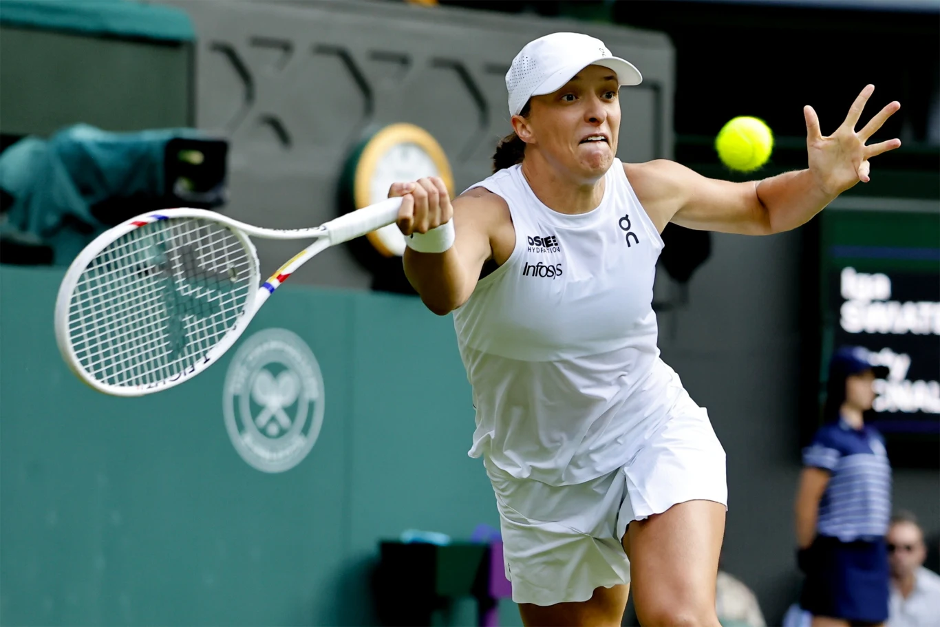 Tenisistka w białym stroju wykonuje dynamiczny forehand na korcie trawiastym, w tle widoczne logo turnieju oraz rozmyci sędziowie.