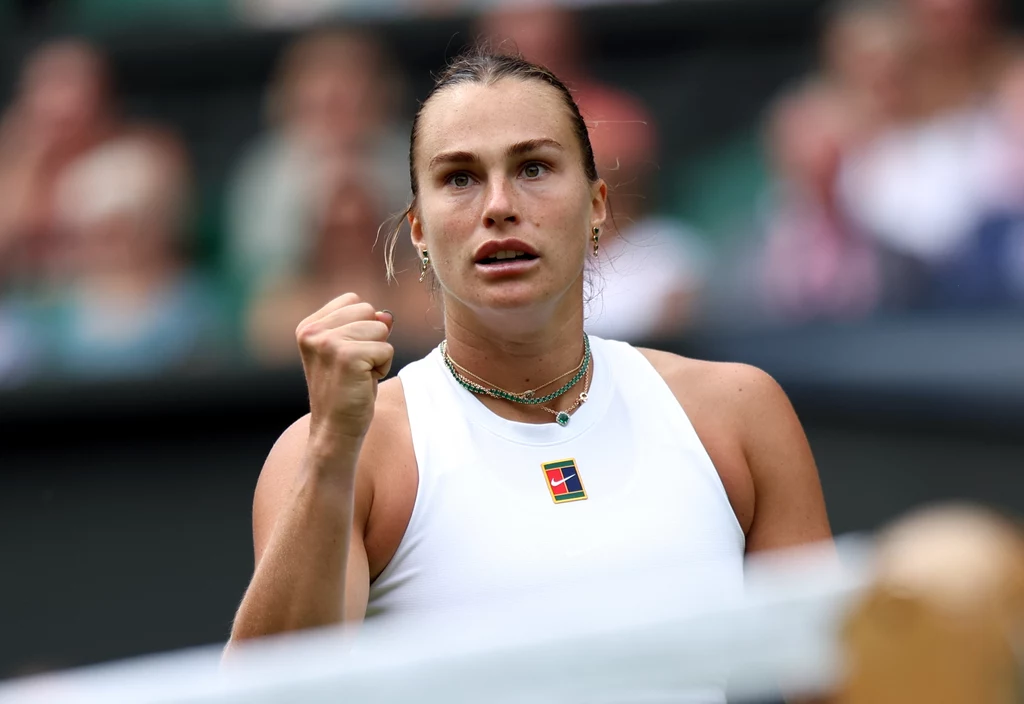 Aryna Sabalenka Tenisistka w białym stroju gestykuluje zaciśniętą pięścią, wyrażając emocje i determinację na korcie tenisowym podczas meczu.