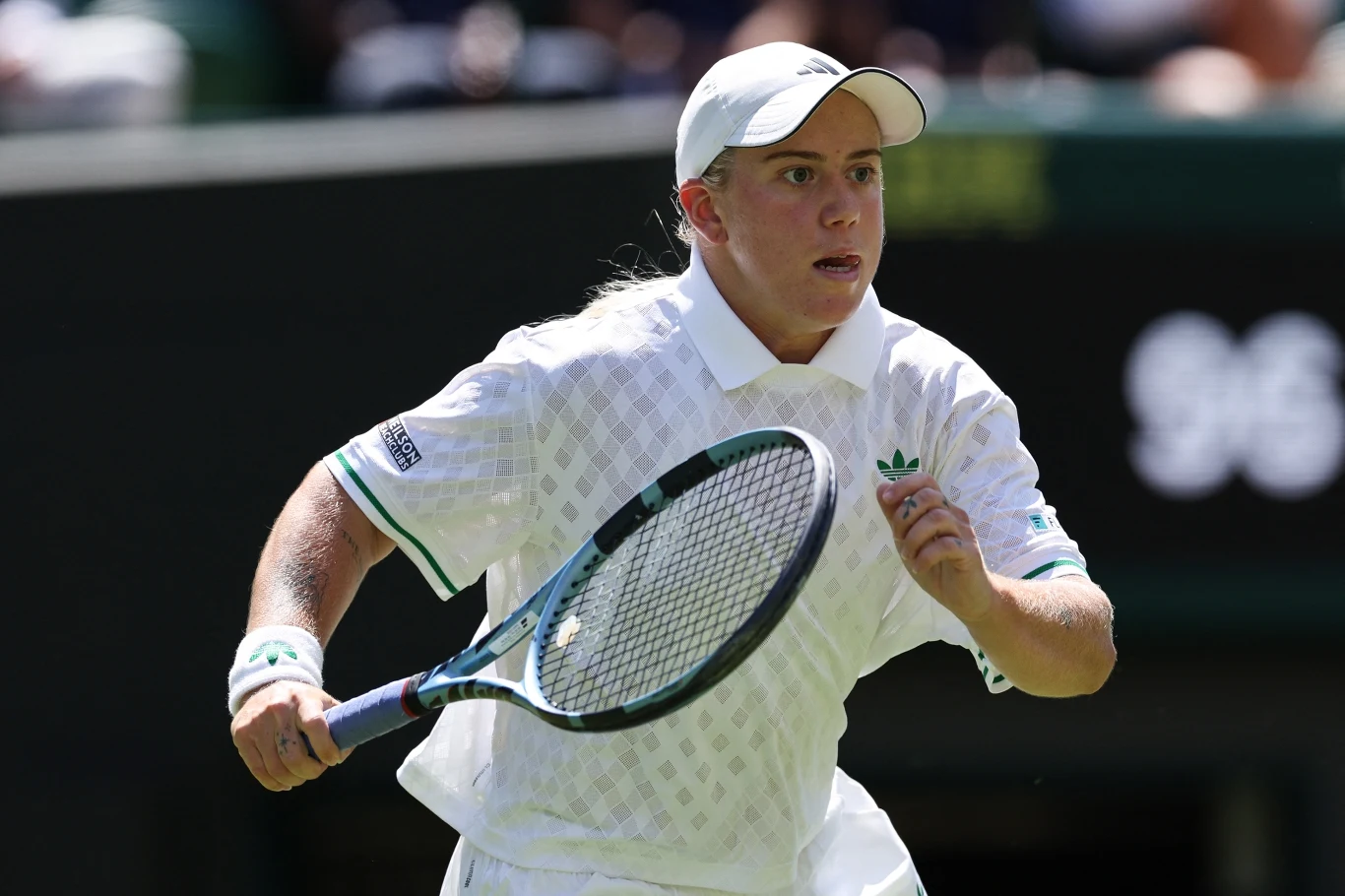 Tenisistka w białym stroju sportowym, z rakietą w dłoni, w ruchu na korcie podczas meczu tenisowego, pełna koncentracja na twarzy, jasne oświetlenie oraz rozmazane tło trybun.