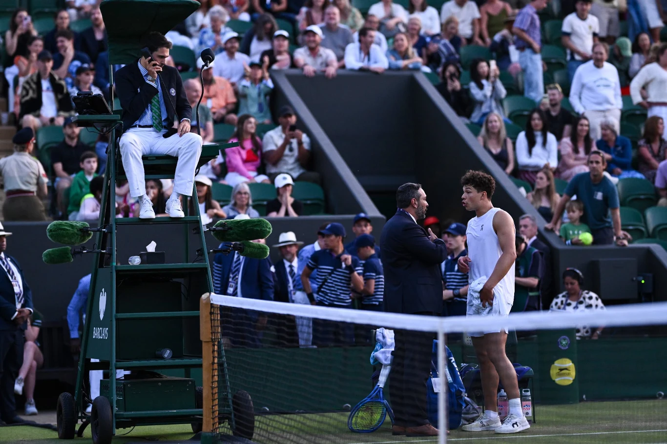 Ben Shelton podczas meczu 2. rundy Wimbledonu Kort tenisowy w trakcie meczu, zawodnik w białym stroju stoi przy siatce rozmawiając z sędzią i inną osobą, sędzia na podwyższeniu obserwuje sytuację, wokół zgromadzona publiczność.