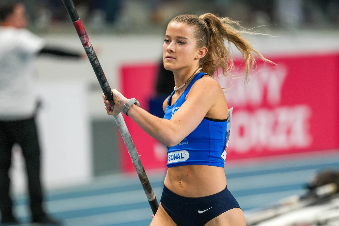 Zawodniczka lekkoatletyczna w stroju sportowym przygotowuje się do skoku o tyczce podczas zawodów halowych; w tle widać innych uczestników i fragmenty trybun.