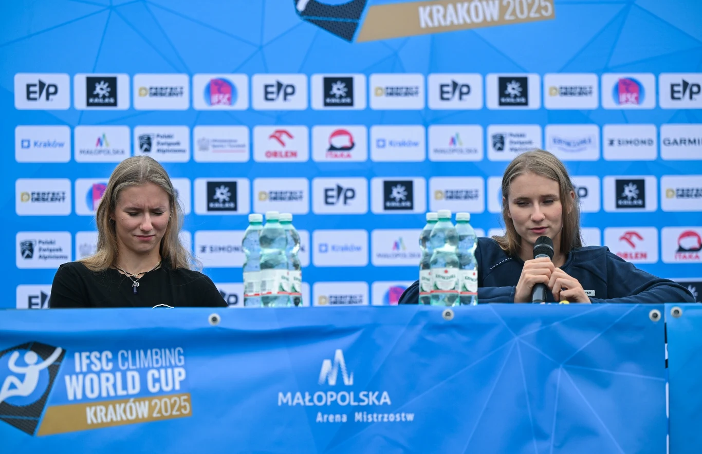 Dwie kobiety siedzące za stołem podczas konferencji prasowej, na tle ściany z logotypami sponsorów i organizatorów IFSC Climbing World Cup Kraków 2025; jedna z nich mówi do mikrofonu, na stole butelki z wodą mineralną.