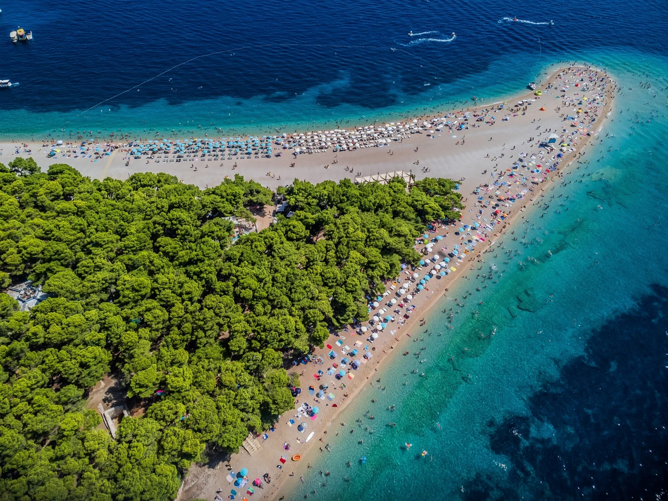 Chorwacja jest często wybierana przez Polaków na miejsce wakacyjnego odpoczynku. Na zdjęciu plaża Zlatni rat na wyspie Brač Plaża o charakterystycznym, wąskim kształcie otoczona przez krystalicznie czyste, turkusowe morze oraz gęsty las sosnowy, liczne kolorowe parasole i tłum wypoczywających ludzi.