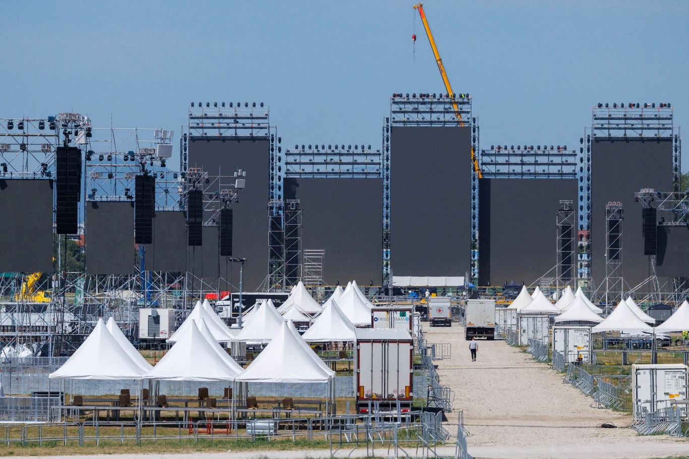 Trwa budowa sceny w Zagrzebiu na koncert grupy Thompson Rzędy białych namiotów ustawione są na placu, w tle widoczna potężna konstrukcja sceniczna z dużymi ekranami LED i reflektorami, na prawo żółty dźwig, a całość otoczona metalowymi ogrodzeniami.