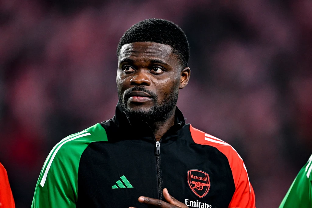 Thomas Partey Piłkarz w stroju treningowym klubu Arsenal z charakterystycznym logo na piersi, stoi na tle rozmytego stadionu, jego wyraz twarzy jest poważny i skupiony.