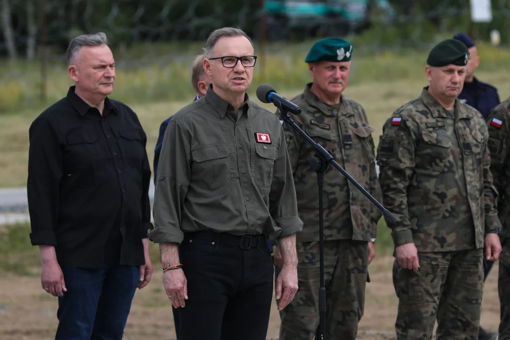 Prezydent Andrzej Duda na granicy polsko-białoruskiej Grupa mężczyzn stoi na zewnątrz, jeden z nich przemawia do mikrofonu ubrany w zieloną koszulę z naszywką, obok stoją żołnierze w mundurach wojskowych z polskimi flagami na rękawach.