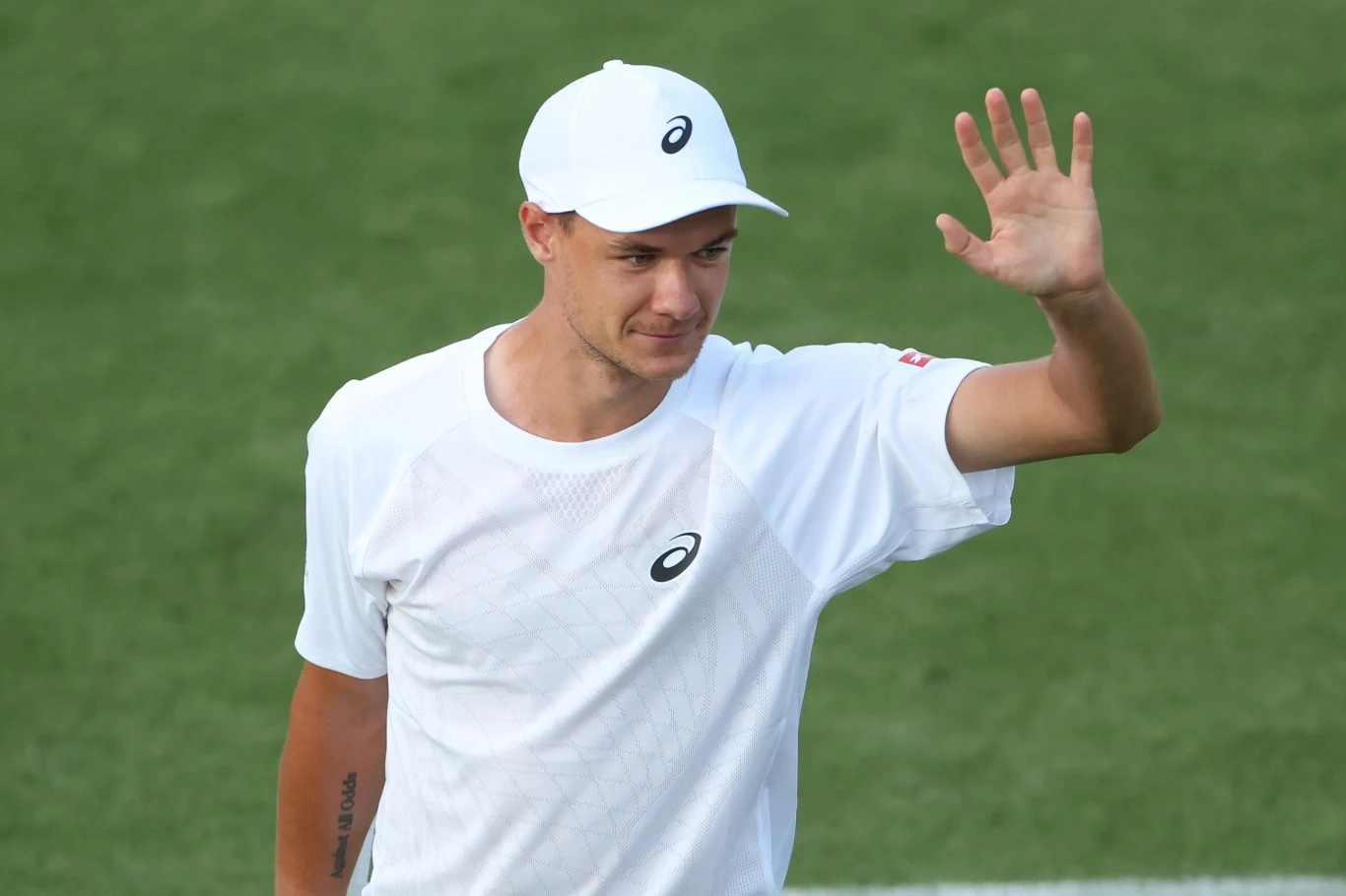Kamil Majchrzak rywalizował z Arthurem Rinderknechem o awans do czwartej rundy Wimbledonu Mężczyzna w białym stroju sportowym i czapeczce marki Asics stoi na zielonym korcie trawiastym oraz unosi dłoń w geście powitania lub pozdrowienia.