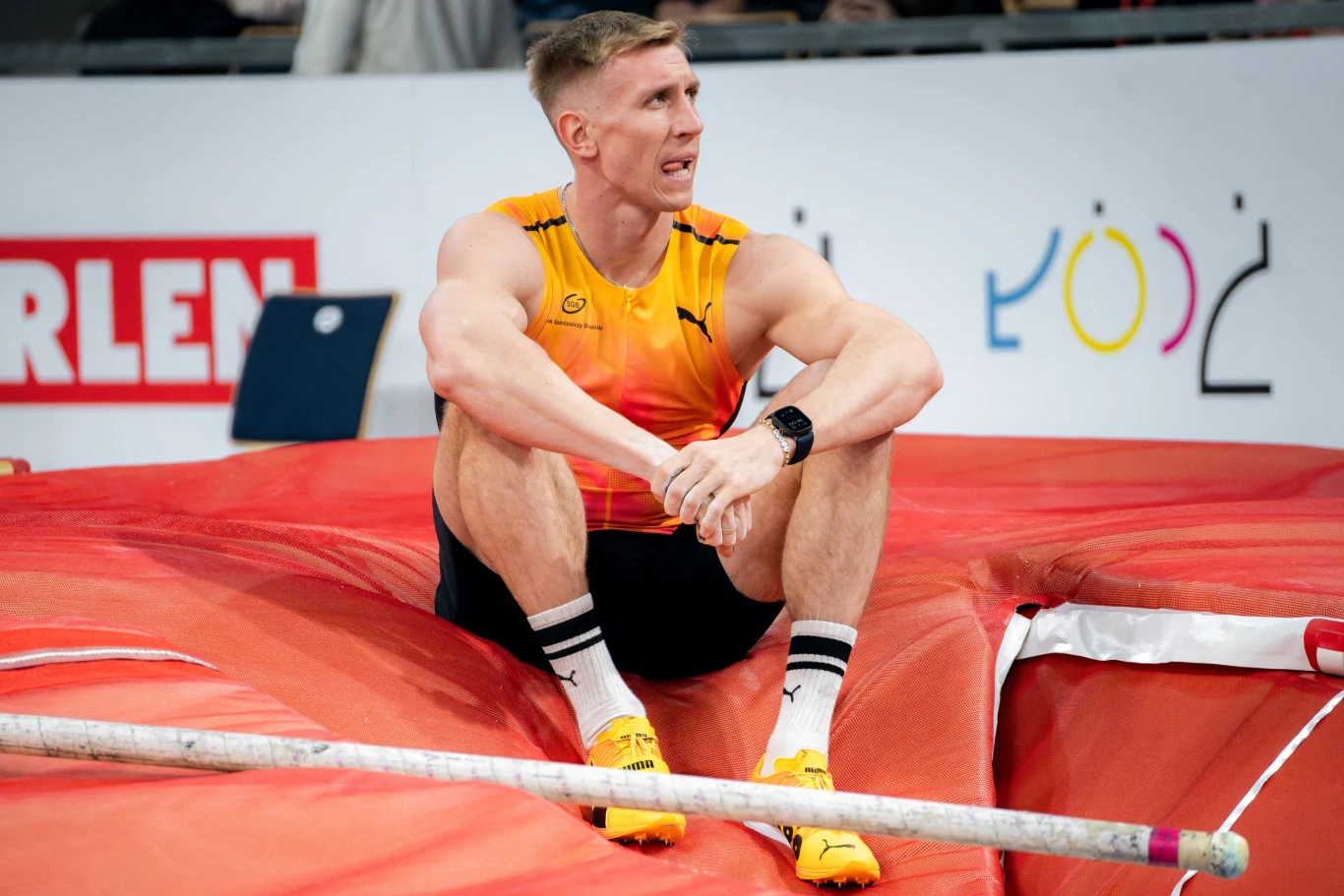 Sportowiec w stroju lekkoatletycznym siedzi na materacu w otoczeniu sprzętu do skoku o tyczce, w tle widoczne elementy hali sportowej i fragmenty reklam.