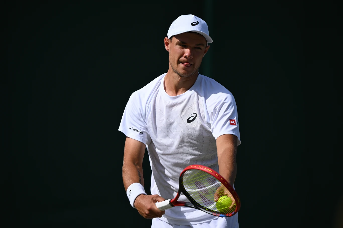 Kamil Majchrzak Tenisista w białym stroju trzymający rakietę i piłkę tenisową przygotowuje się do serwisu, ciemne tło podkreśla sportową sylwetkę i skupienie zawodnika.