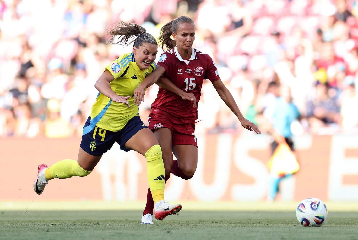 Mecz Szwecja - Dania, Euro 2025 Dwie piłkarki rywalizujące o piłkę na boisku, jedna w żółtym stroju, druga w bordowym, dynamiczna akcja podczas meczu piłki nożnej, w tle rozmyta publiczność.