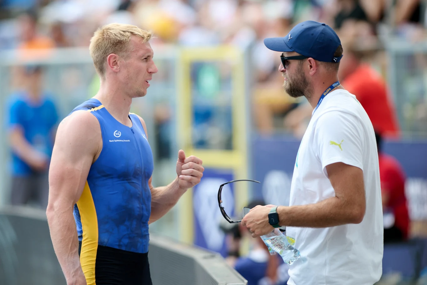 Piotr Lisek i Marcin Szczepański Sportowiec w stroju zawodniczym rozmawia z trenerem trzymającym butelkę wody i okulary przeciwsłoneczne na tle stadionu sportowego. Obaj są skupieni na rozmowie, w tle widać rozmytą publiczność.