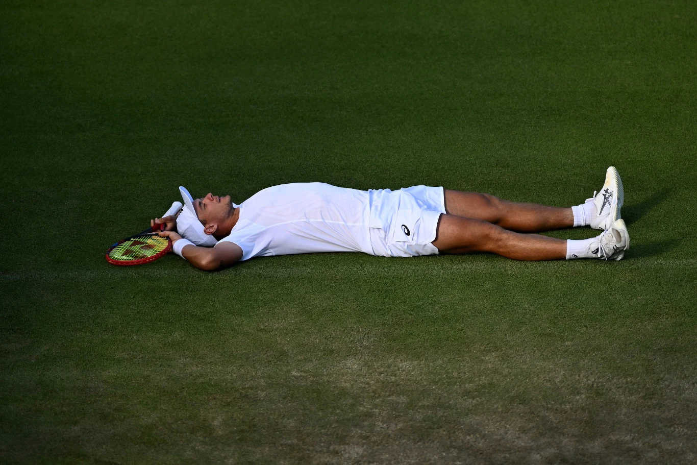 Kamil Majchrzak po ostatniej piłce w meczu trzeciej rundy Wimbledonu położył się na korcie. Tak przeżywał swój awans Sportowiec leży wyczerpany na trawie kortu tenisowego, ubraną w biały strój i opaskę na głowie, z rakietą tenisową w dłoni. Oświetlenie wskazuje na ważny moment zakończenia meczu.