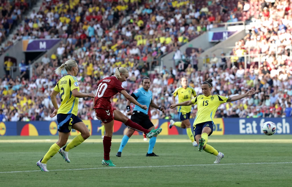 Pernille Harder w meczu ze Szwecją na Euro 2025 04.07.2025 Piłkarka w czerwonym stroju wykonuje strzał na bramkę, blokowana przez dwie zawodniczki w żółtych koszulkach, wokół których stoi sędzia oraz tłum widzów na trybunach.